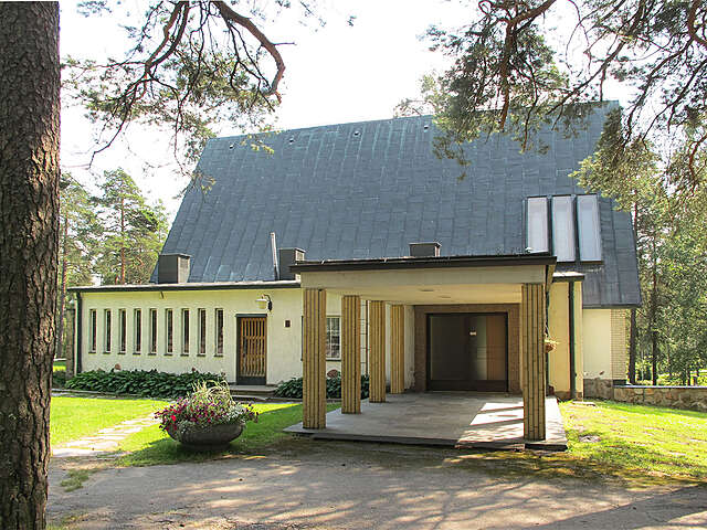 Chapel exterior with a colonnade in front of the entrance, white walls and a black gable roof surrounded by pine trees.
