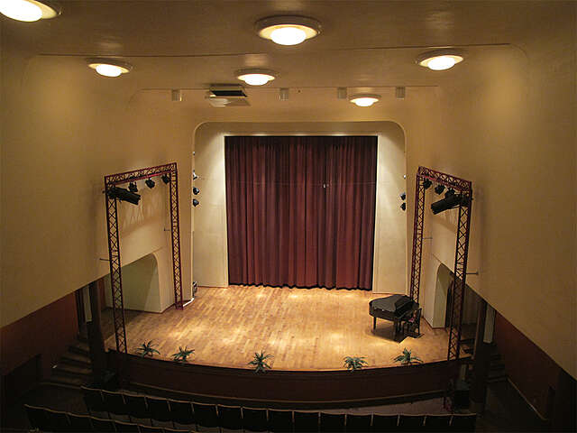Stage with red curtains, a wooden floor, a grand piano and white walls.