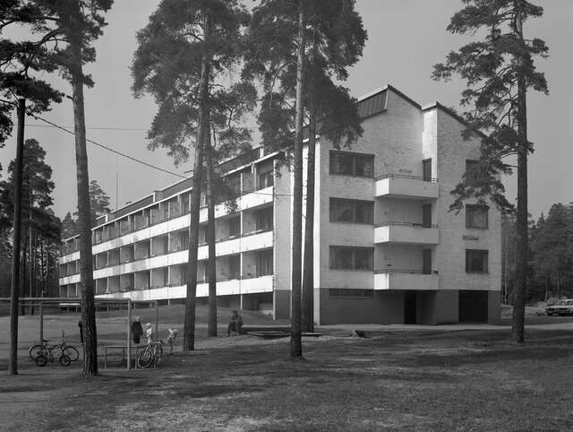 Three-storey apartment block behind a yard and pines