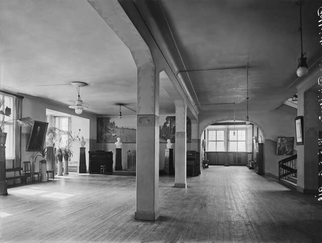 School interior with two columns, statues and paintings distributed along the walls and a bench in the left corner.