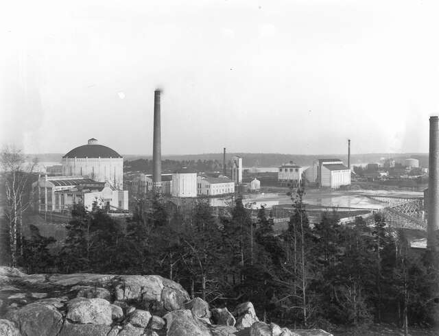 Industrial buildings in the horizont, rocky and forested landscape in the foreground