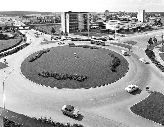 Black and white picture of a roundabout with old cars in it and commercial buildings in the background.