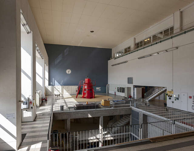 Image of the machine hall with bright red machinery, that stands out from the deep blue shaded wall at the back of the space. Natural, direct sunlight flows in from the high vertical windows.