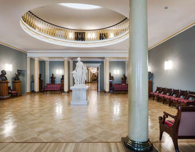 Large waiting room with wooden chairs, sculptures, grey walls and a balcony below the window ceiling.