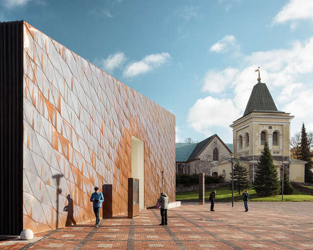 Closed copper facade with an opening in the middle standing on a paved square, medieval church and belfry in the background