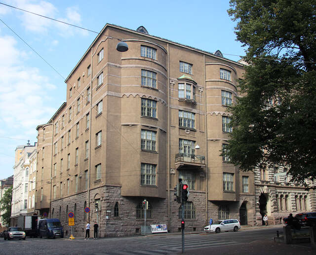 Building with a rubble ground floor and elegantly formed recessed entrances that conceal fine copper-clad doors. tHe upper levels of the building are covered in beige plaster and there are some decorative lines at the second highest floor.
