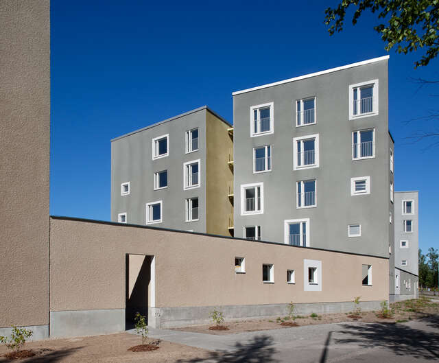Two five-storey grey block houses rising behind a beige-coloured wall with an entrance and square holes