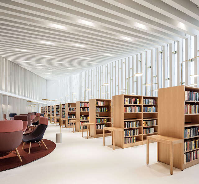 White space with vertical battens in the window wall and joists, wooden bookshelves, burgundy-coloured armchairs