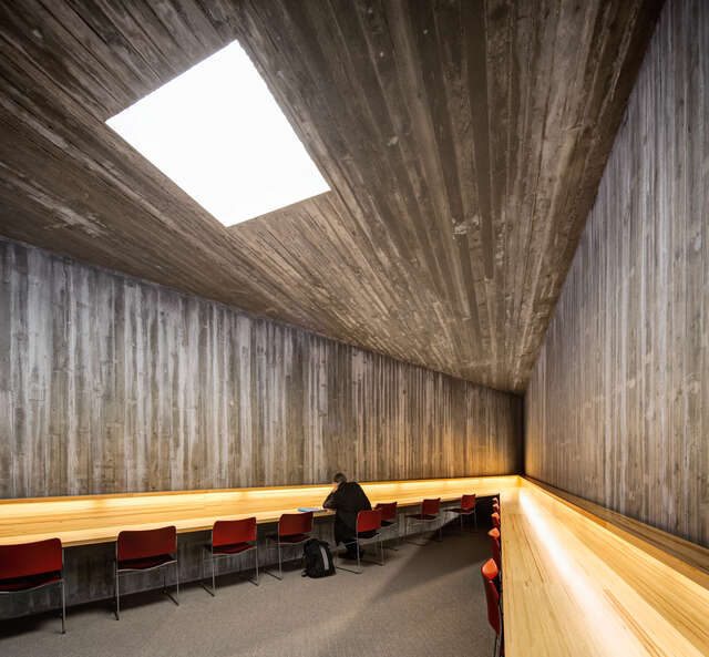 A person reading at a wooden long table on a red chair, surrounded by concrete walls and ceiling