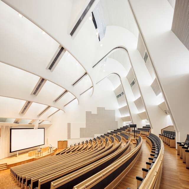 Large lecture hall with wooden floors and seating structures, white walls and a tall curved ceiling.