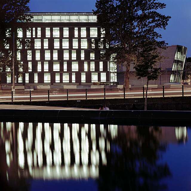 The lights of a grey building with offset windows reflect on the water.