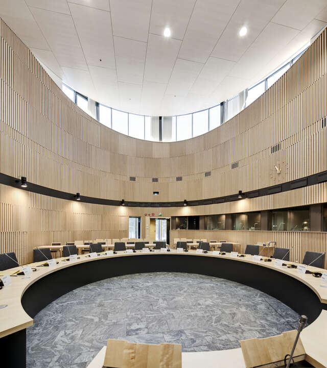 A space with curvy, wooden walls. Inside the space, there is a large oval shaped table and plenty of chairs around it.