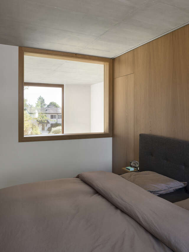 A room with a small square window and a large bed.