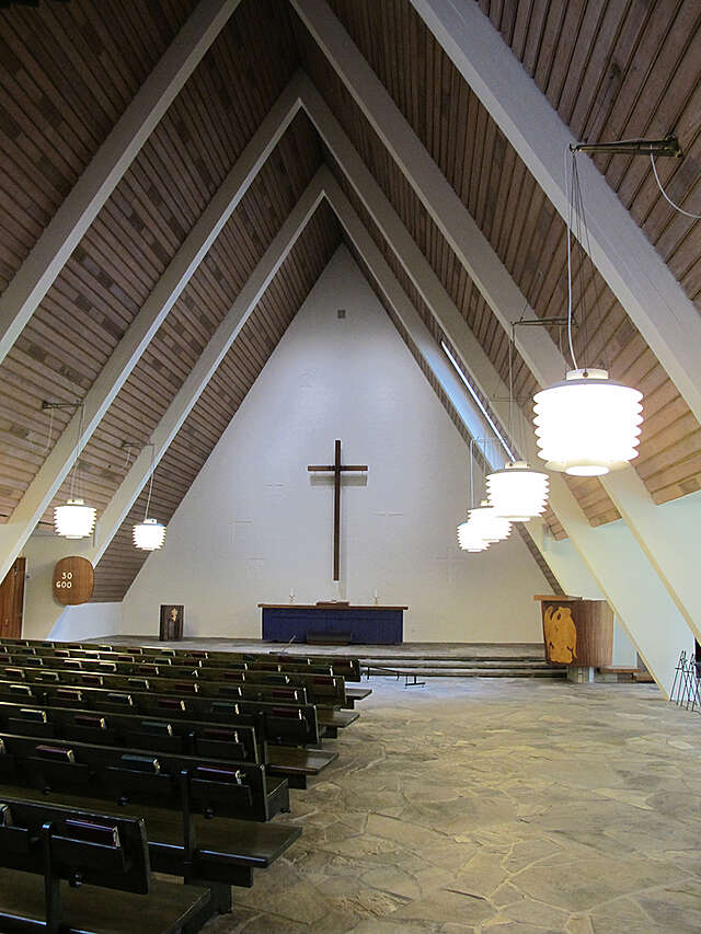 View to the altar, black bench rows on the left, hanging light fixtures on the walls and wooden paneling on the gable roof.