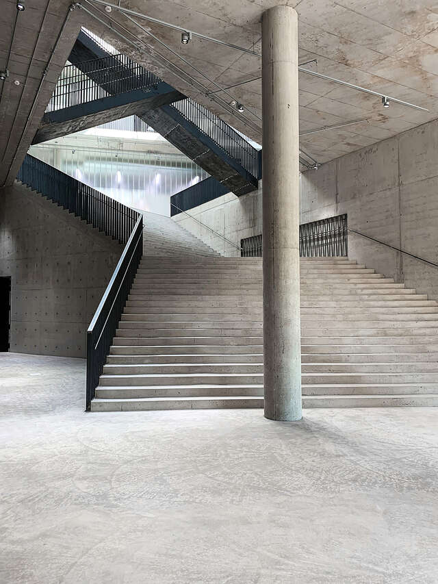Interior of concrete structure with large concrete staircase. One large round concrete pillar in the center of the image.
