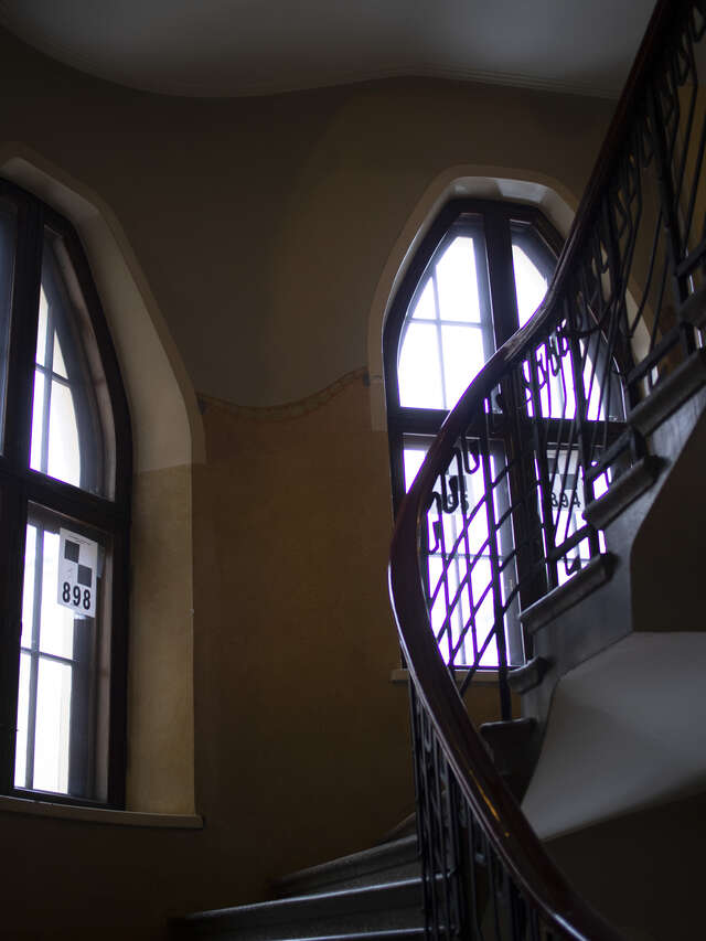 Stone staircase with a black ornate railing in a terra-cotta colored hallway with ornate windows.