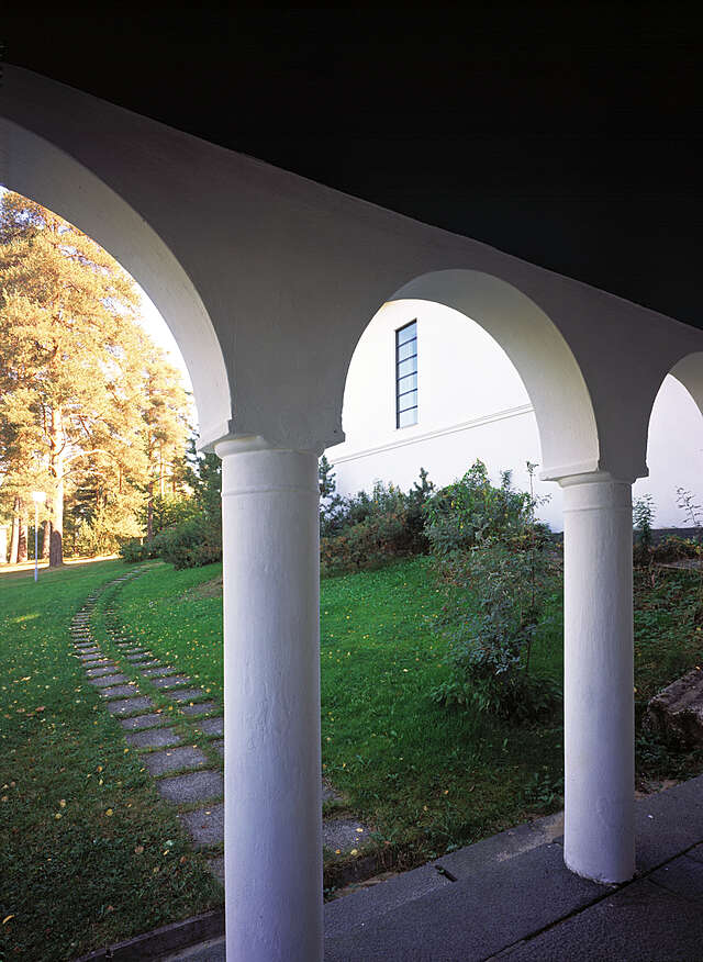 White pillars supporting arches at the exterior of the building, the yard and tall pine trees in the background.