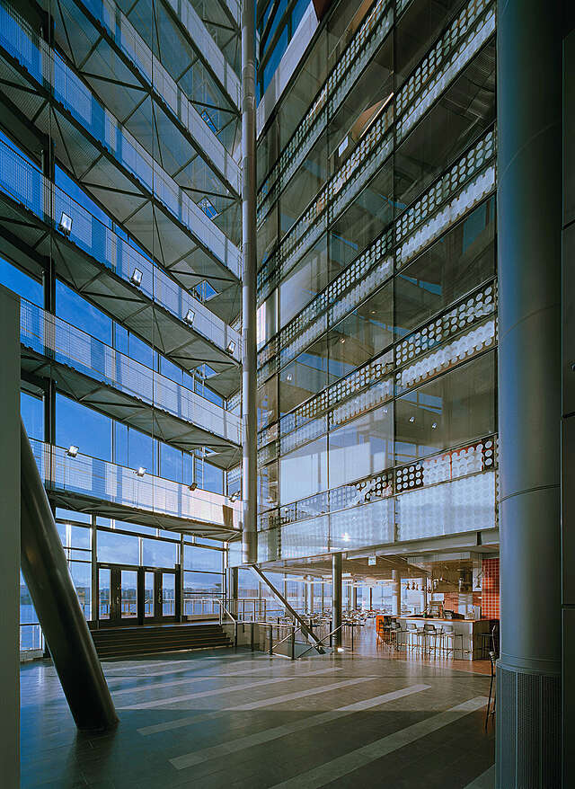 Lobby with a very high ceiling and large glass walls. Ib the back there is a cafeteria.