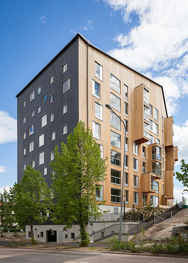 Apartment building with a wooden exterior and gable roof, around the building there are many trees.