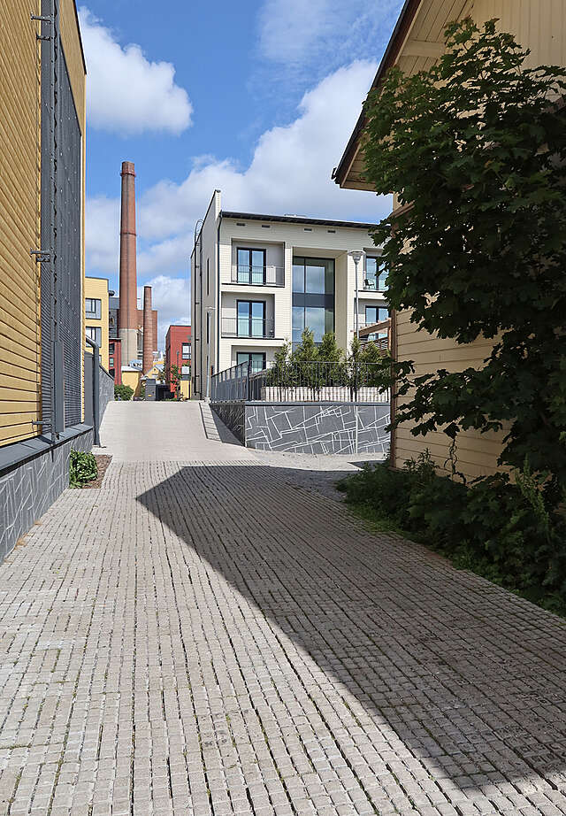 Pedestrian street between a new, yellow block of flats and an older, white wooden house. Factory chimneys in the distance.