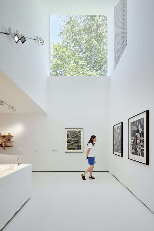 White interior of an art gallery with a window high up on the wall. One person on ground level looking at artworks.