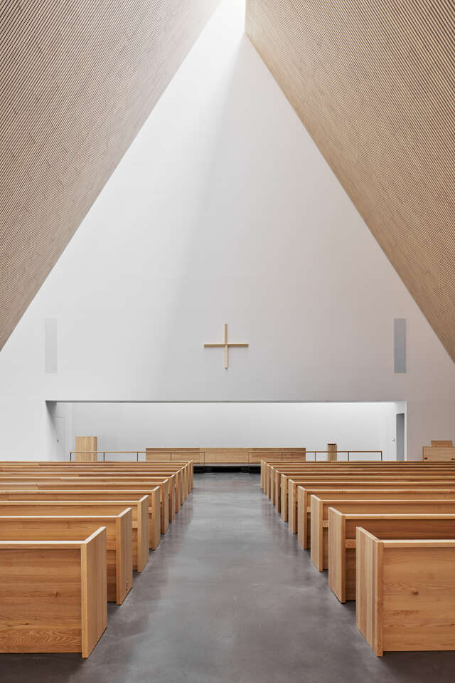 Steep wooden ceiling, white wall with wooden cross on it and wooden church pews.