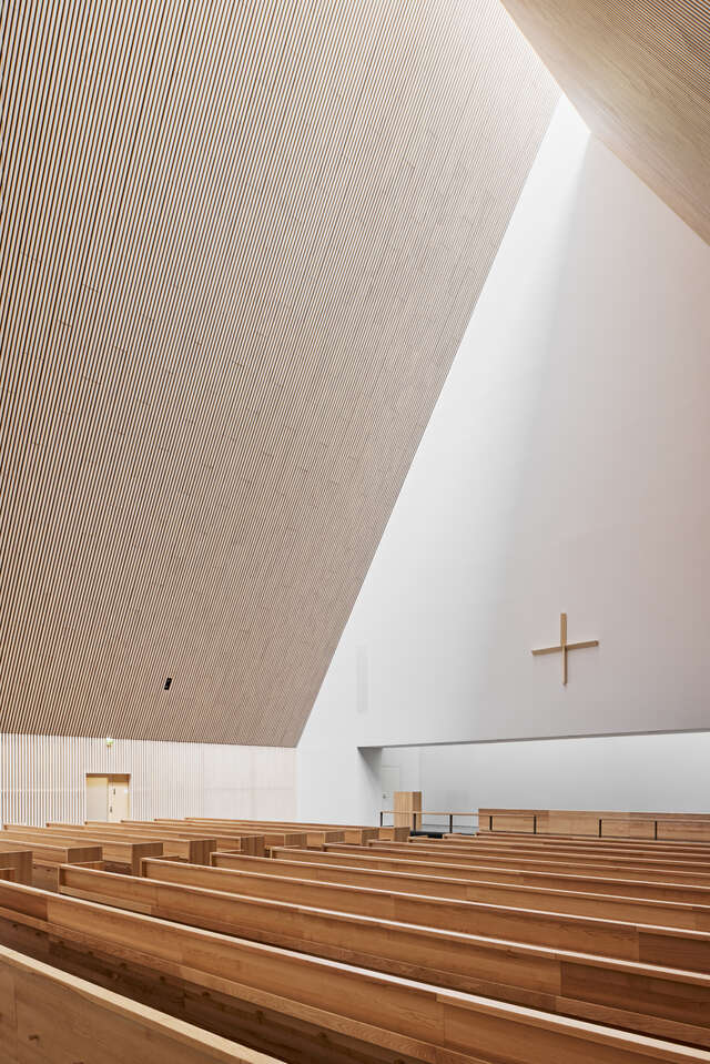 Steep wooden ceiling, white wall with wooden cross on it and wooden church pews.