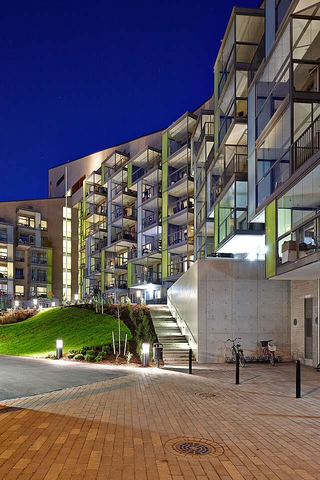 Multiple story residential building with protruding balconies with colored green and blue glass details in evening illumination.