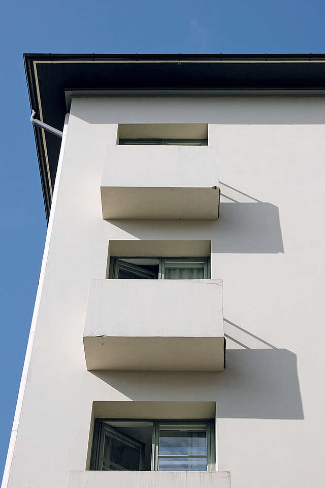 Simple white building with rectangular protruding balconies and a black roof.