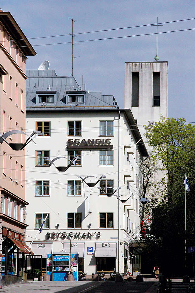 White hotel building in the city, an ice cream shop and streetlights in the front, a church tower in the back.