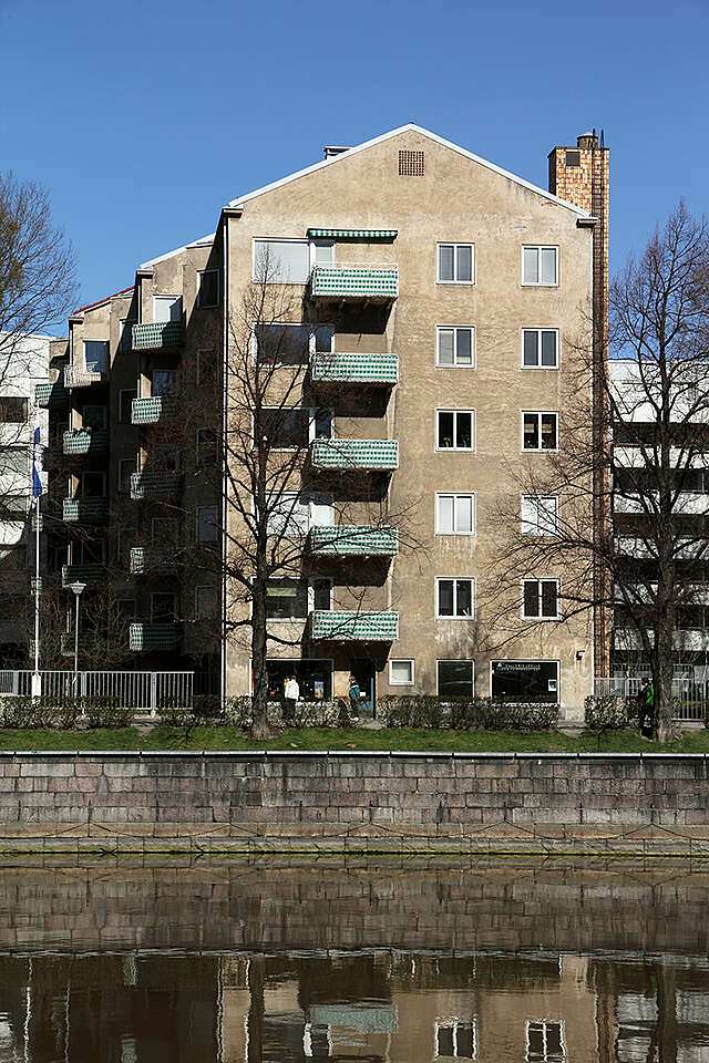 Multiple story building in front of a river, with a grey plaster facade and balconies with white metal railings covered from the inside with a green and white striped fabric.