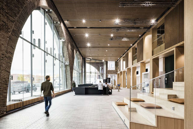 Sisäkuva aulasta, vasemmalla suuri tiilikaari, jossa ikkuna, oikealla puiset istumaportaat, ihmisiä